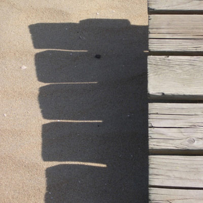 De uiteinden van de planken van het houten vlonder werpen scherpe schaduwen op het zand.